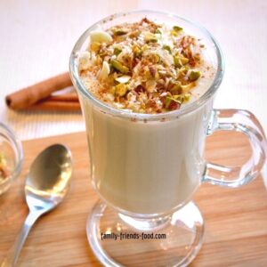 Glass cup of sahlab topped with nuts and cinnamon, on a small wooden board with a teaspoon and a dish of nuts.