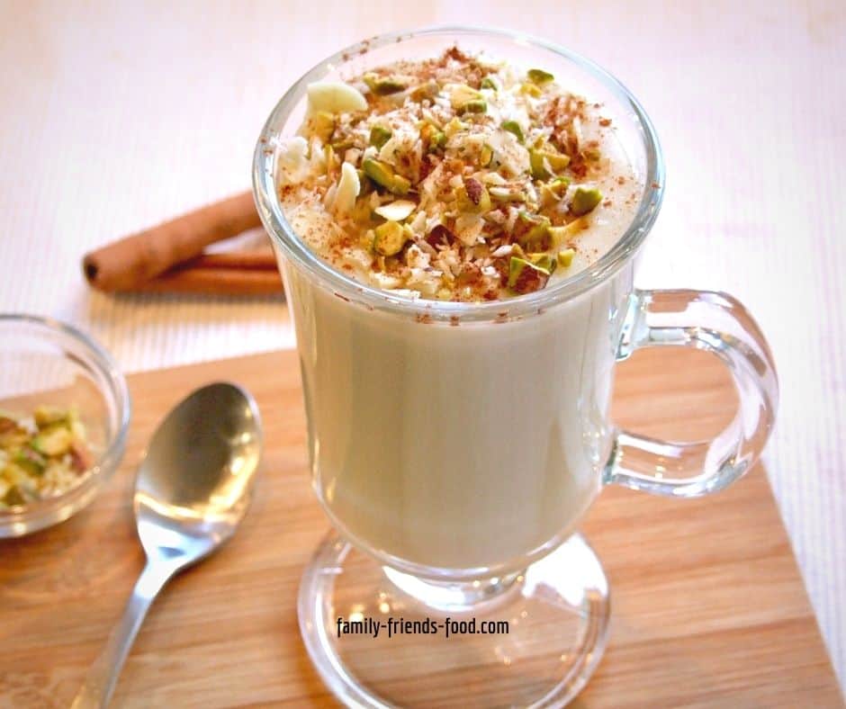 Glass cup of sahlab topped with nuts and cinnamon, on a small wooden board with a teaspoon and a dish of nuts. 