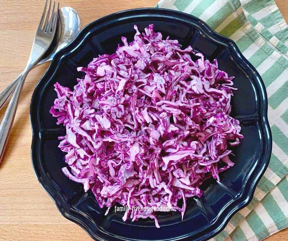 A bowl of red cabbage salad, on a wooden table, with a serving fork and spoon.