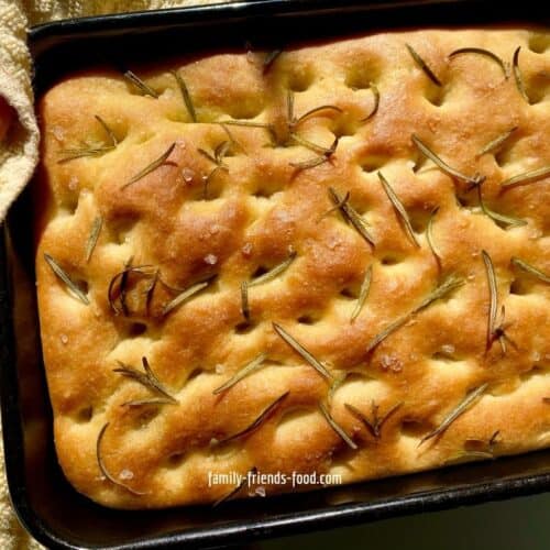 Focaccia in a tin, hot from the oven, held on the left by a hand with a yellow cloth.
