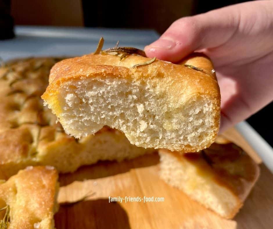 A child's hand holding a piece of focaccia, the rest of the focaccia is in the background.