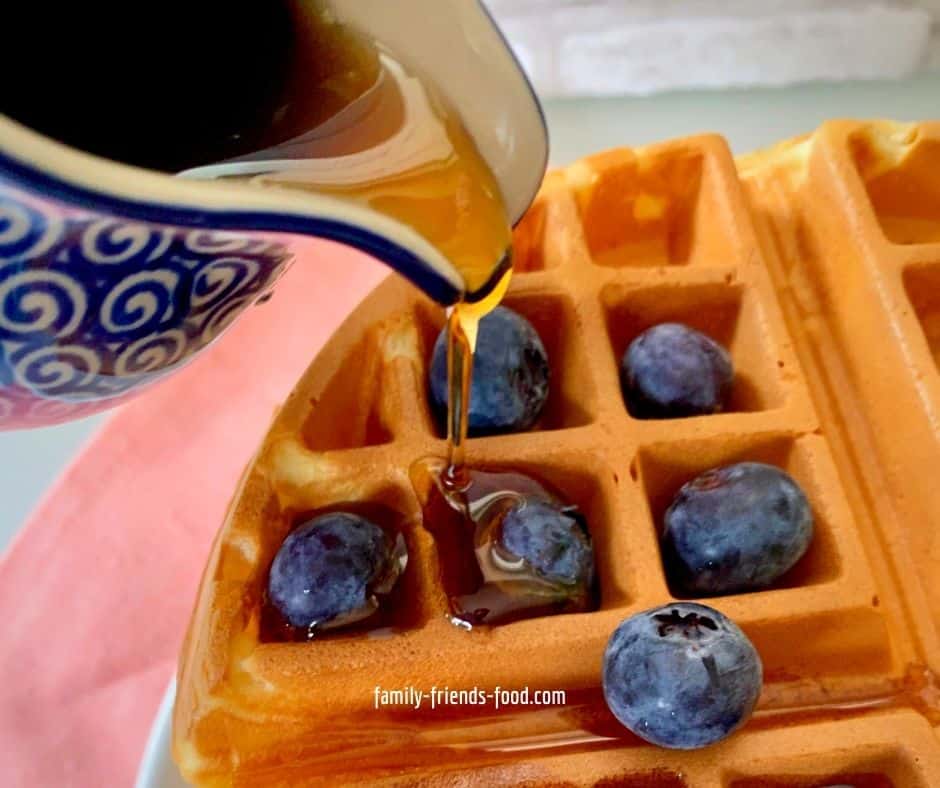 Close up image of a blue patterned jug pouring maple syrup over a waffle with blueberries.