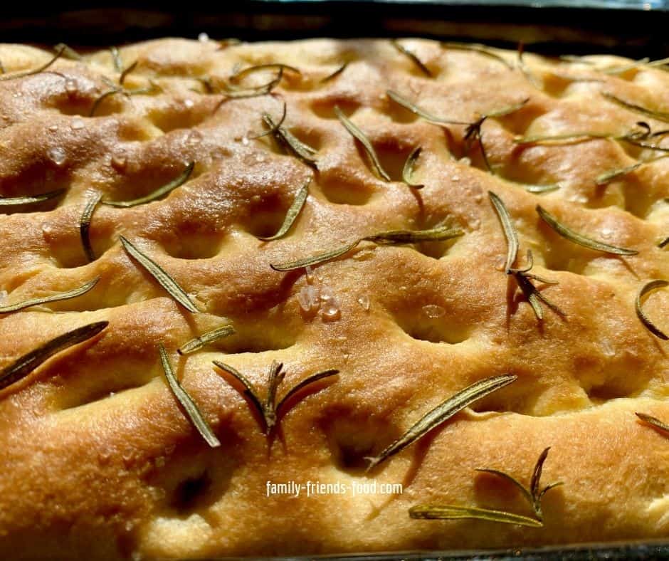 Close up of the surface of focaccia showing dimpled bread with grains of sea salt and leaves of rosemary.