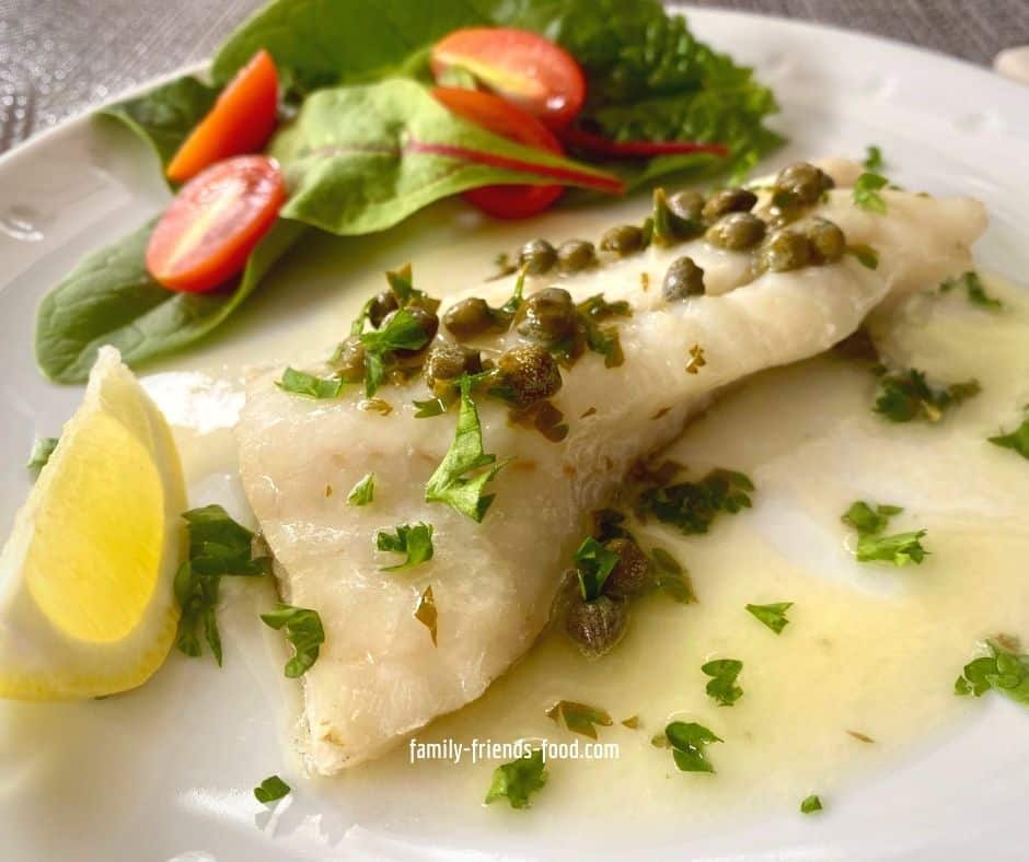 Close up image of a piece of white fish with lemon caper butter sauce, sprinkled with chopped parsley, on a white plate. Salad leaves and tomatoes are also on the plate.