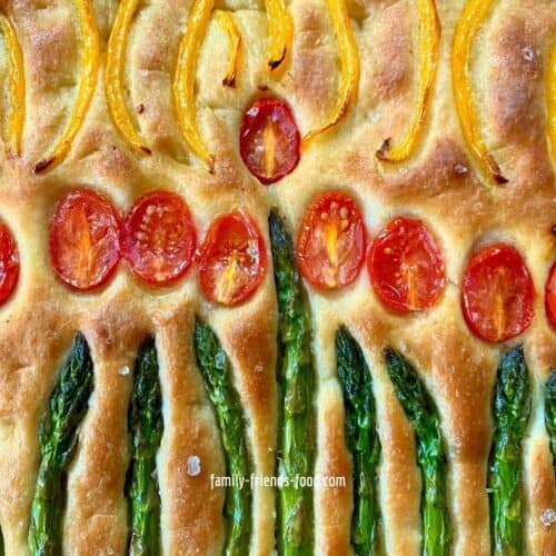 Close up image of the surface of a baked Chanukah focaccia, showing asparagus spears, halved baby plum tomatoes, and yellow pepper slices, arranged into the shape of a Chanukiah with glowing candles.