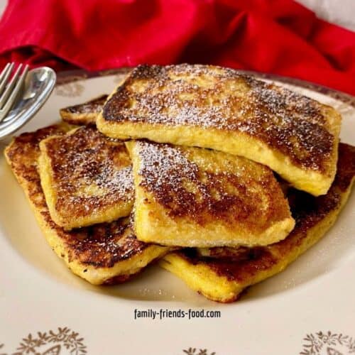 A stack of French toast slices on a fancy plate with spoon and fork, red napkin in background.