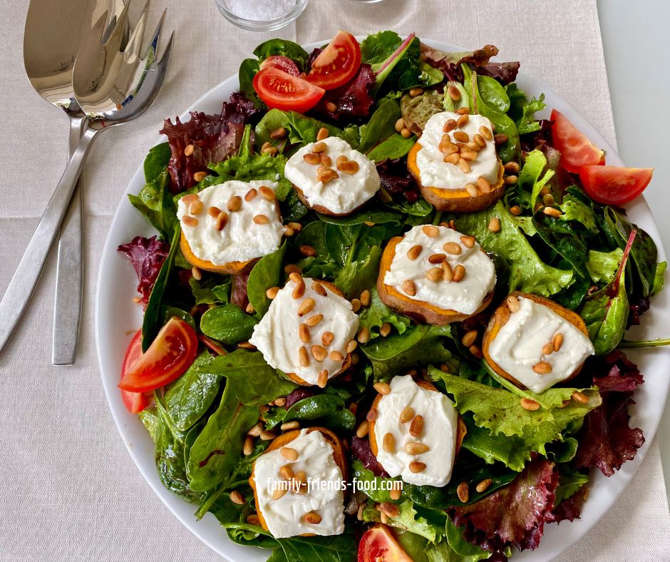 A platter of dressed salad leaves and slices of goats-cheese-topped sweet potato, sprinkled with pine nuts. Wedges of tomato are arranged at intervals around the edge of the platter. The platter is on a cream cloth, salad servers to the left.