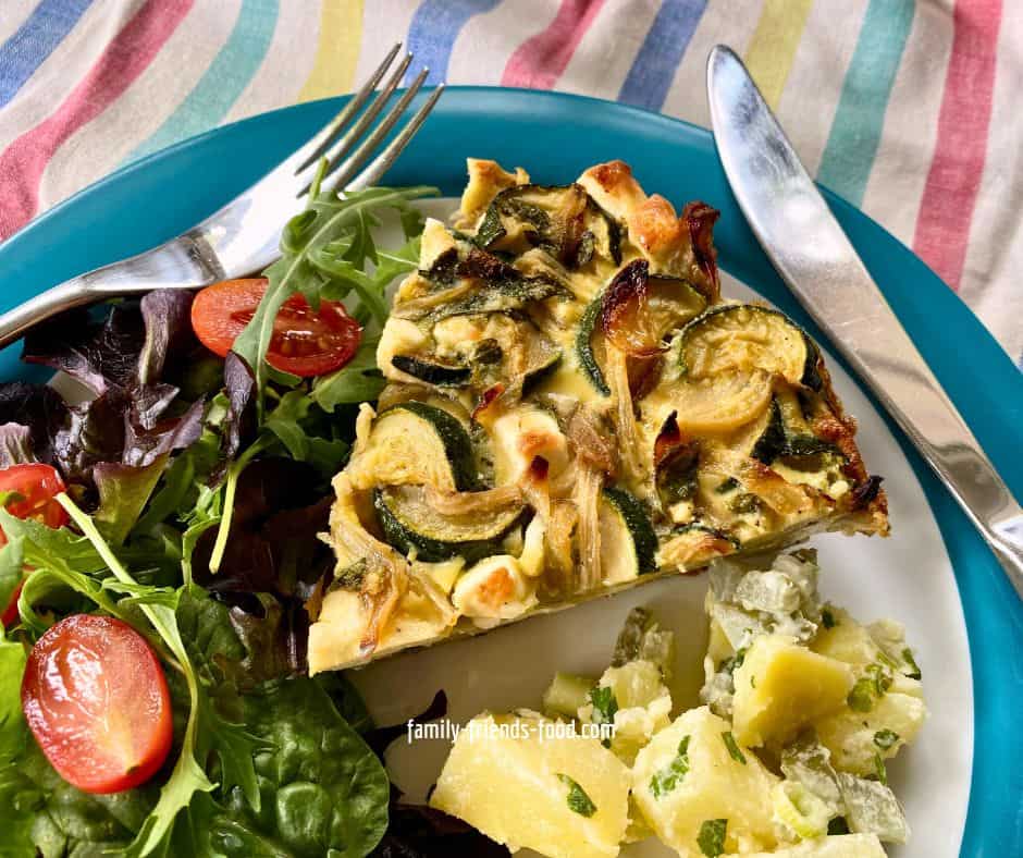 Close up image of a slice of courgette feta and mint pashtida on a plate with leafy salad and potato salad, with a knife and fork, and a striped cloth underneath the plate.