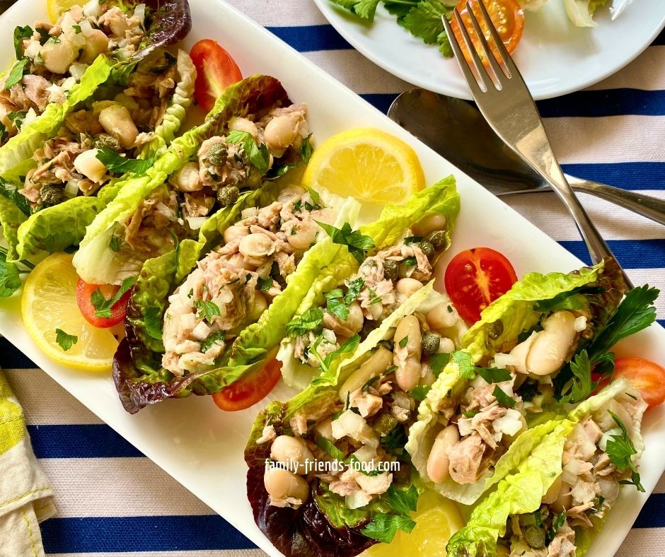 Overhead image of a platter of Italian tuna and beans salad served in lettuce cups, and another small plate of the same, sitting on a blue and white striped cloth, with a fork.