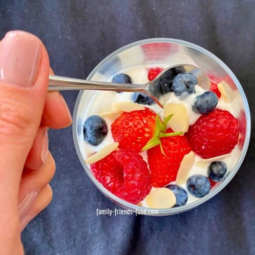 Overhead image of a hand holding a spoon, taking a spoonful of fruit and nut yogurt parfait. The topping of mixed berries and flaked almonds over a layer of creamy yogurt is visible in the glass.