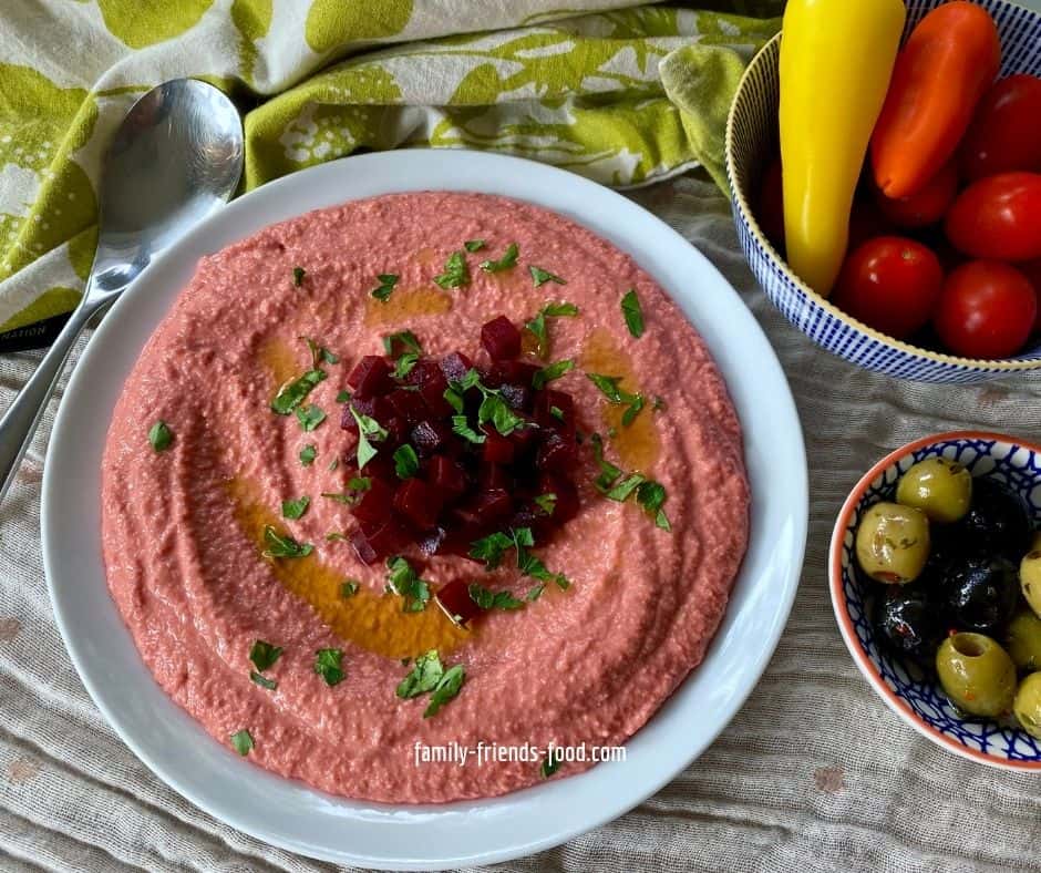 A plate of beetroot hummus, topped with diced beets, olive oil and chopped parsley. On the right are a dish of olives and a dish of small peppers and tomatoes.