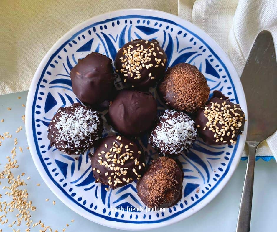 Overhead image of chocolate-coated tahini date truffles, some plain, some sprinkled with sesame seeds, some with desiccated coconut and some with cocoa powder, on a blue and white patterned plate. The plate is partly resting on a cream coloured cloth with blue edging, and part of a serving utensil can be seen to the right. Sesame seeds are scattered on the tabletop beside the plate.