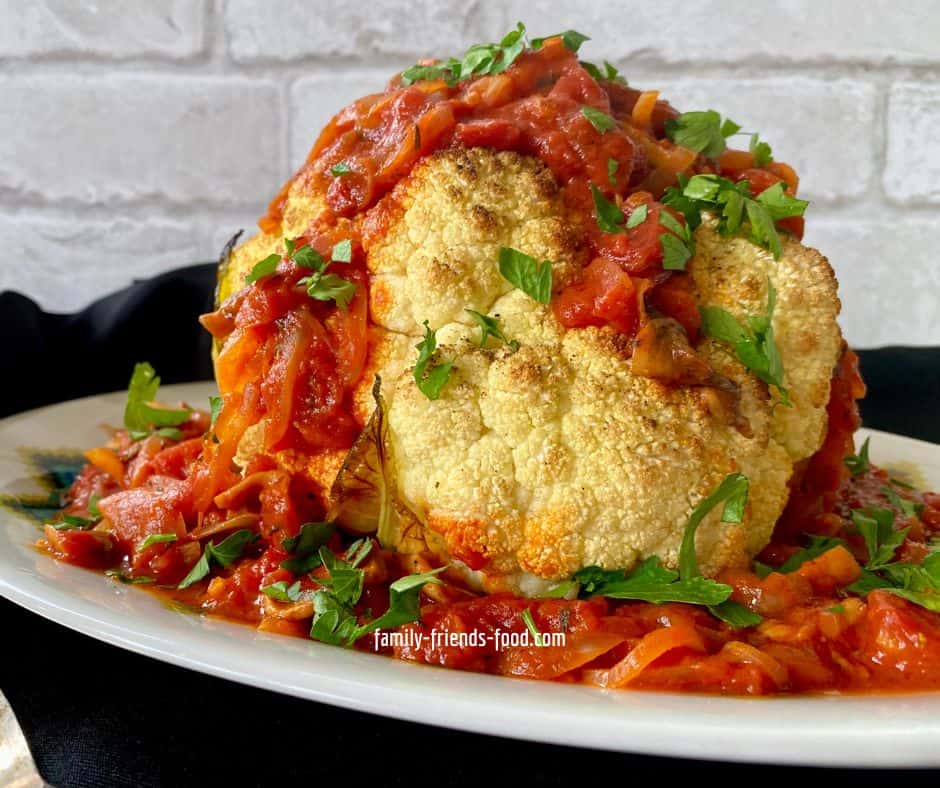 A whole roasted cauliflower on a china serving platter, surrounded by and topped with tomato mushroom sauce, and sprinkled with chopped parsley. The platter sits on a black cloth.