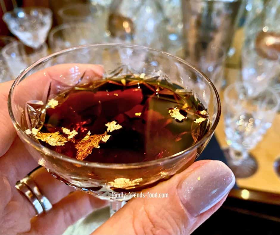 A glass of chanukah cocktail, topped with gold flakes, held aloft in front of an array of vintage glassware.