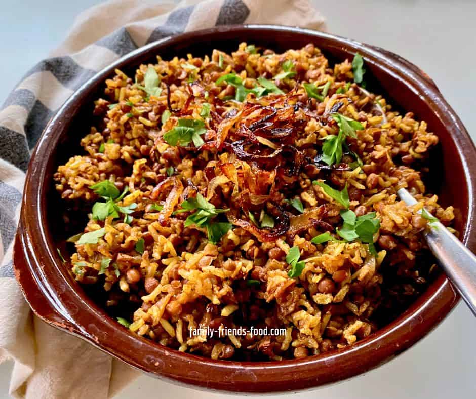 A dish of baked mujadara - rice & lentils - topped with fried onions and a sprinkle of chopped parsley. A spoon is sticking out of the dish, and a black and white cloth is visible to the left.