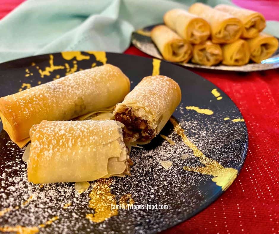 Two Haman's fingers on a plate, dusted with icing sugar. The frontmost one is cut in half and angled to show the fruit and nut filling. A stack of Haman's fingers on another plate, and a light blue napkin, can be seen in the background