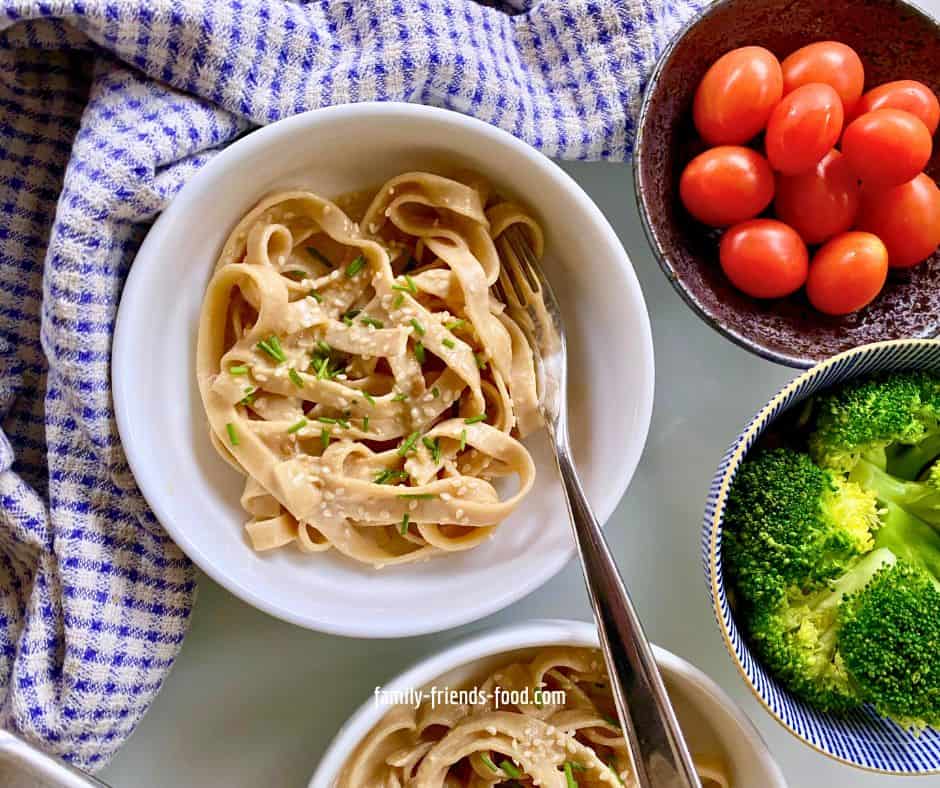 A white china bowl of tagliatelle noodles in miso tahini sauce, sprinkled with sesame seeds and snipped chives, with a fork sticking out. A blue and white cloth is to the left and behind. A second bowl of pasta is just visible in front, and bowls of broccoli and cherry tomatoes can be seen on the right.