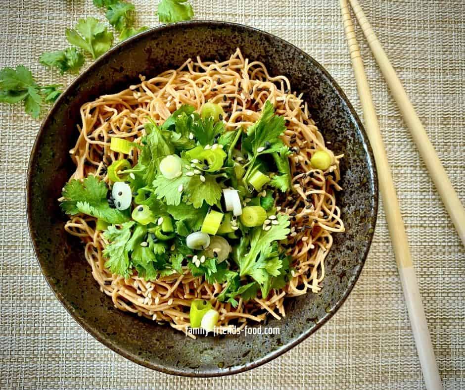 Overhead image of a dark coloured bowl of cold sesame noodles, topped with a generous amount of fresh coriander (cilantro), sliced spring onions (scallions) and a sprinkle of black and white sesame seeds, on a woven mat. Chopsticks are visible to the right of the bowl.
